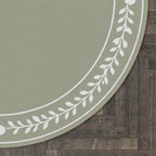 A close-up of a round rug with a sage green center and a white leafy border, shown on a dark wood plank floor.