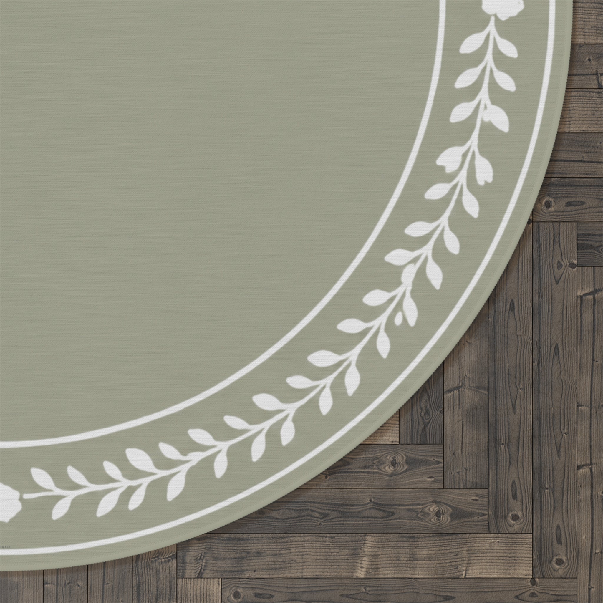A close-up of a round rug with a sage green center and a white leafy border, shown on a dark wood plank floor.