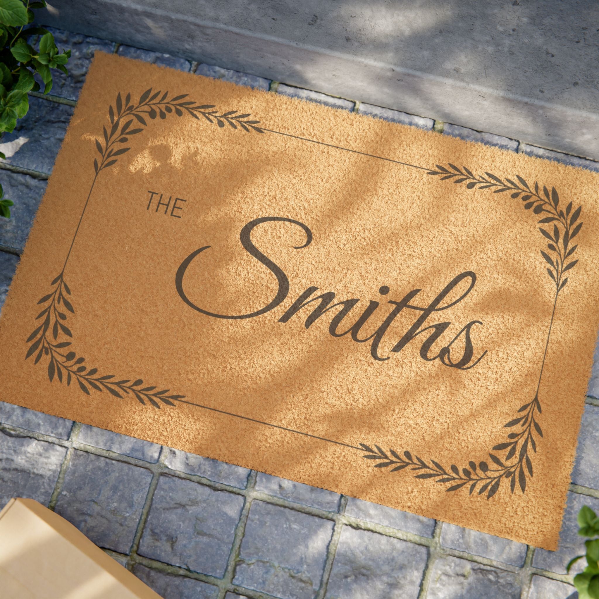 A personalized coir doormat with the name 'The Smiths' in dark brown script, surrounded by a decorative leaf border, placed on a gray stone paver surface.