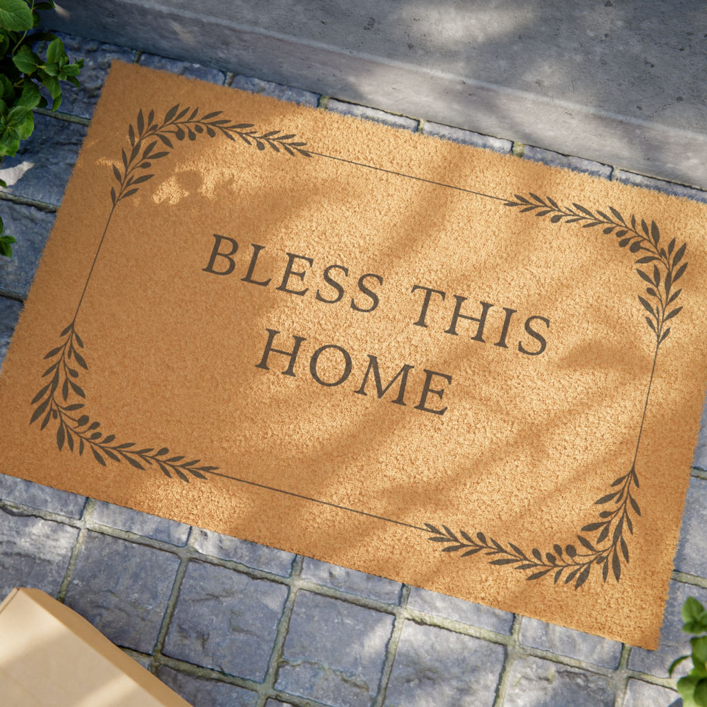 A natural coir doormat with the phrase 'BLESS THIS HOME' in dark lettering, surrounded by a decorative leaf border, placed on a gray stone tile surface.