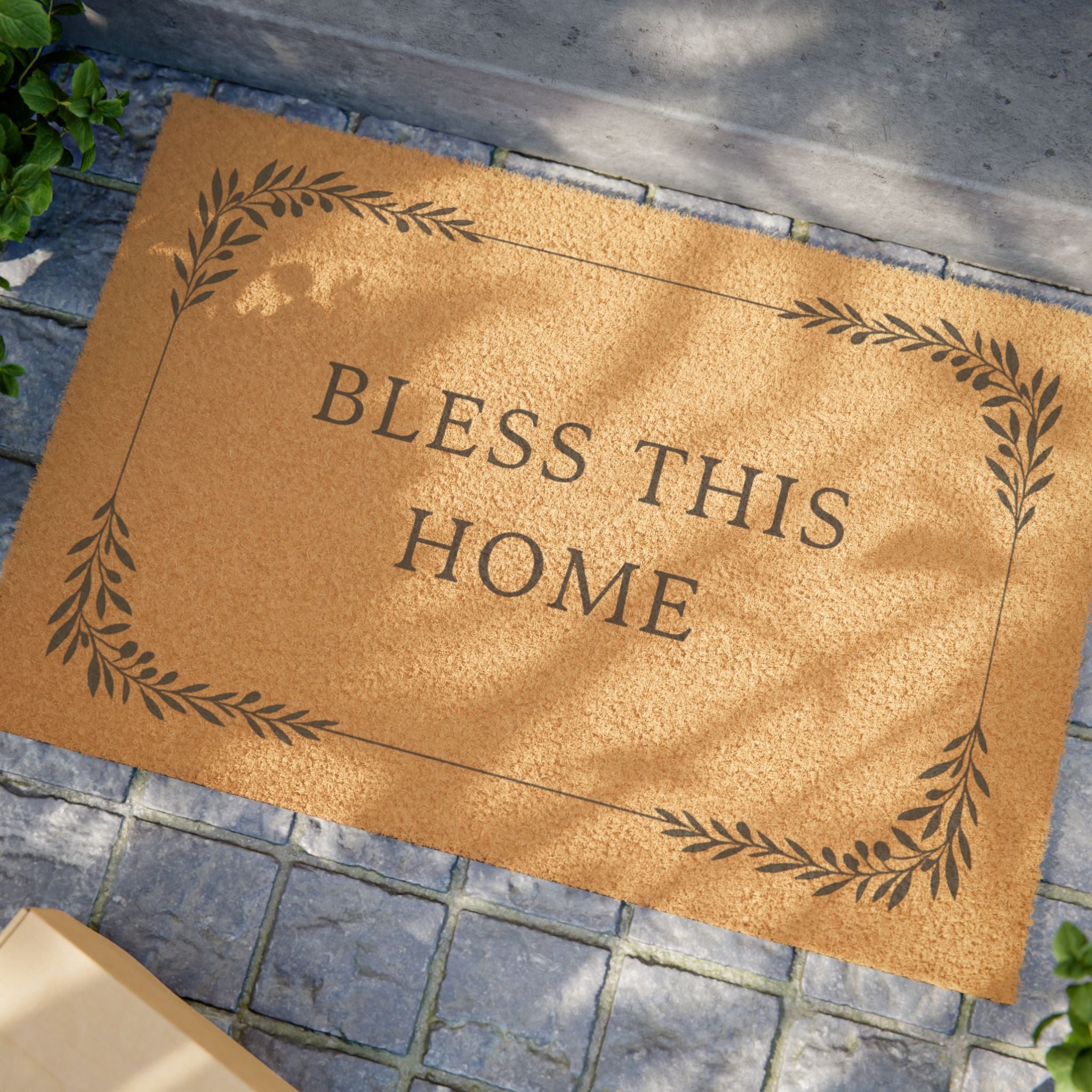 A natural coir doormat with the phrase 'BLESS THIS HOME' in dark lettering, surrounded by a decorative leaf border, placed on a gray stone tile surface.
