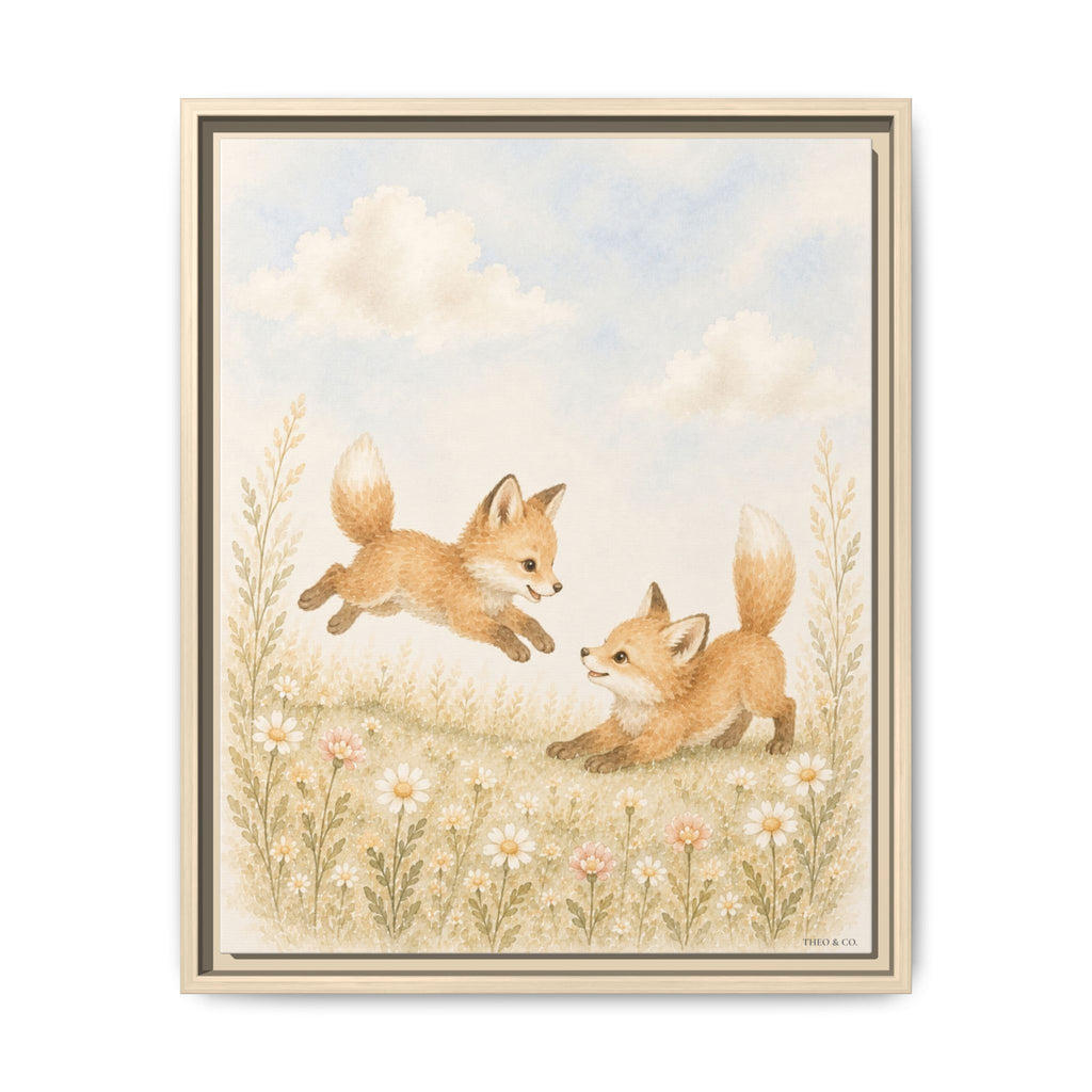 A framed art print featuring an illustration of two playful foxes in a grassy field with wildflowers under a soft blue sky. The print is presented in a light wood frame against a clean white background.
