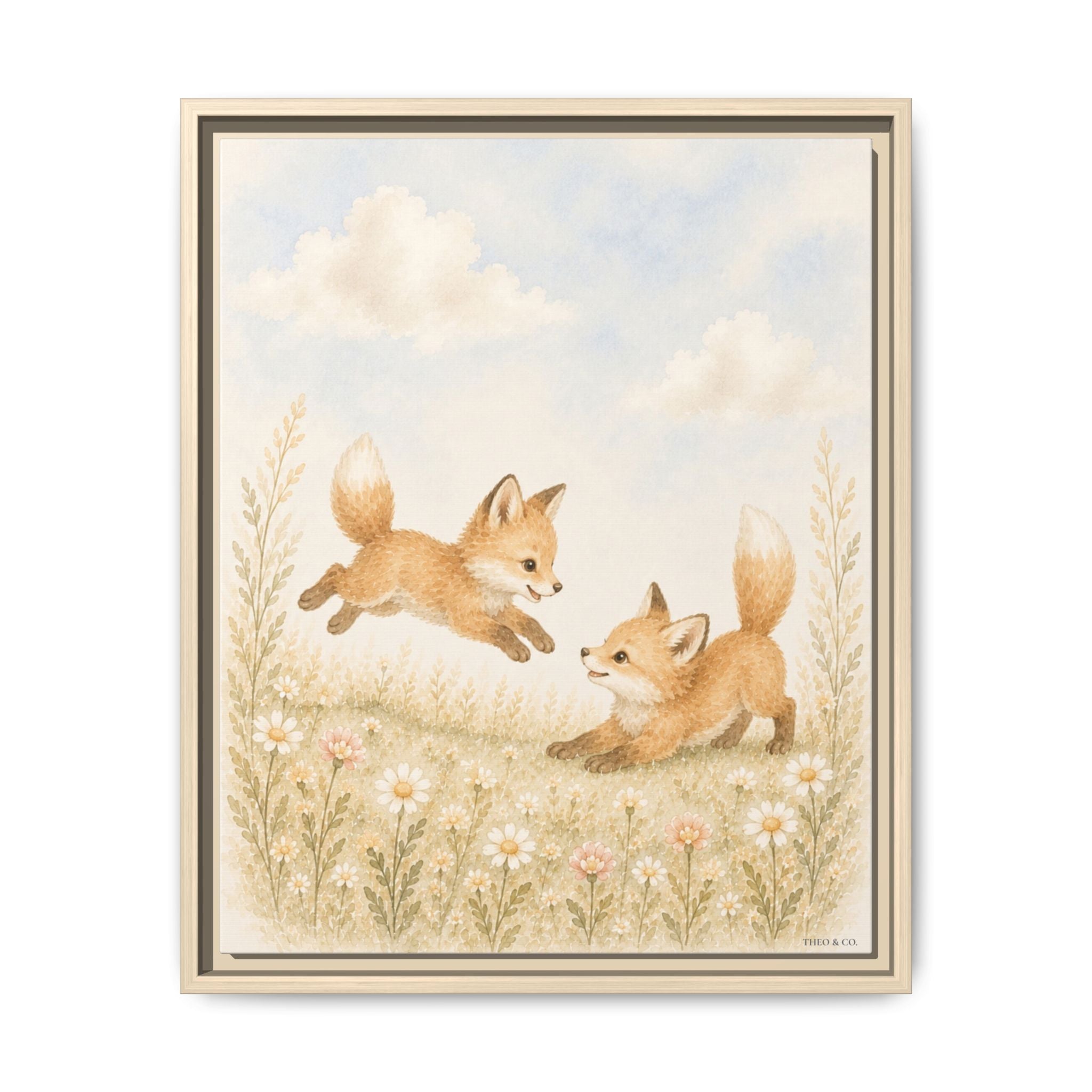 A framed art print featuring an illustration of two playful foxes in a grassy field with wildflowers under a soft blue sky. The print is presented in a light wood frame against a clean white background.