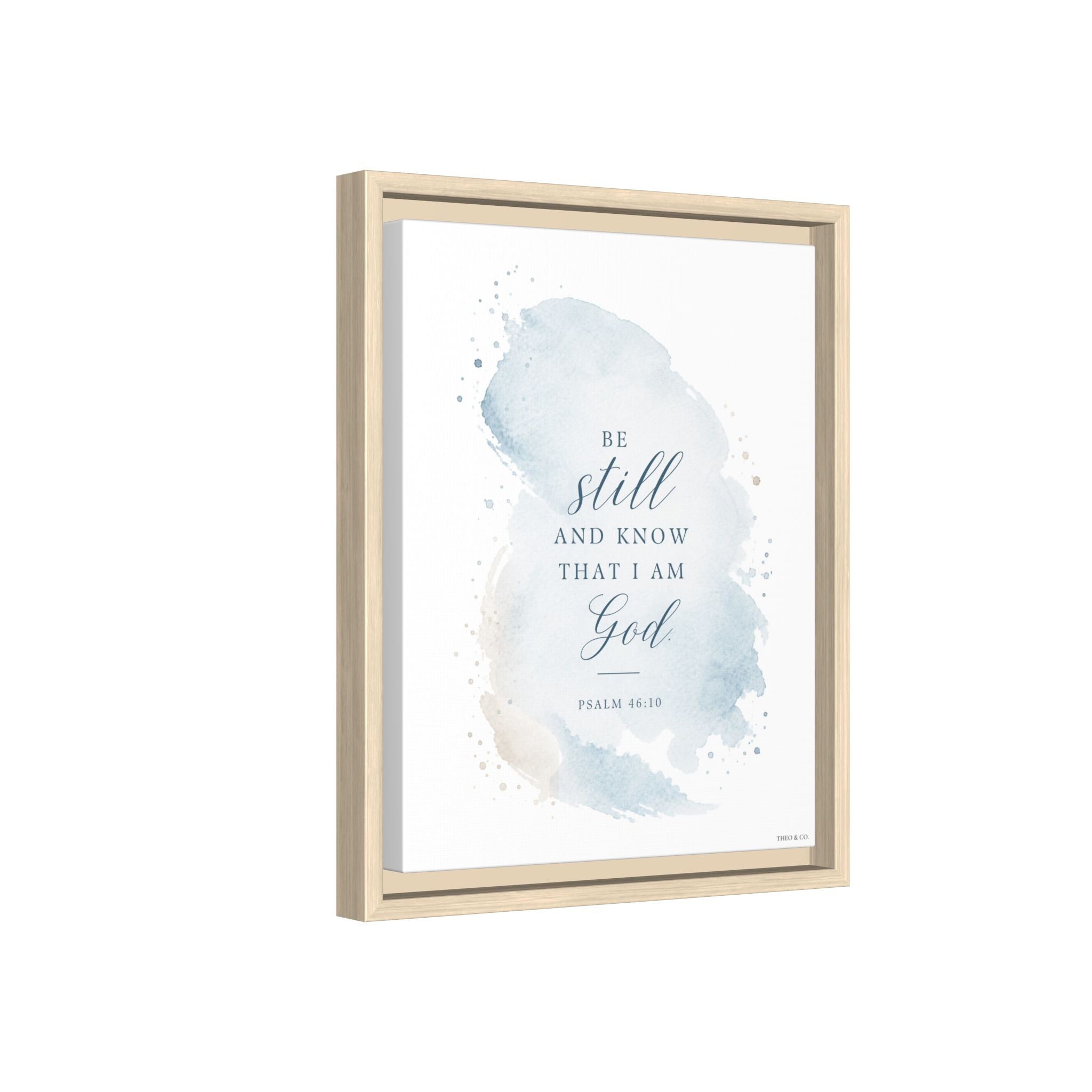 Framed wall art featuring the Bible verse 'Be still and know that I am God' in dark blue script over a soft blue watercolor wash on a white background, shown in a light natural wood frame against a white backdrop.