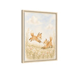 Framed wall art featuring an illustration of two foxes in a meadow under a soft blue sky, shown at a slight angle with a light wood frame on a white background.
