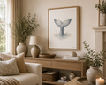 A framed whale tail art print hangs on a beige wall above a wooden console table in a styled living room with plants, a lamp, and a sofa.