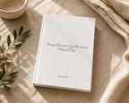 A white hardcover book with the printed message 'Always Remember You Are Loved Mom & Dad' and 'THEO & CO.' on the cover, styled on a beige linen surface with a small bowl, leafy branch, and textured throw blanket.