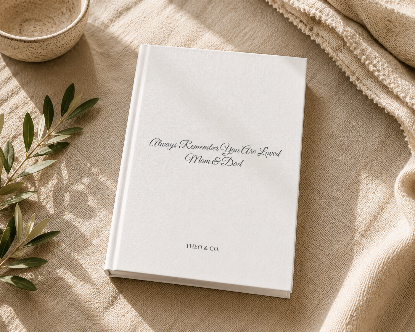 A white hardcover book with the printed message 'Always Remember You Are Loved Mom & Dad' and 'THEO & CO.' on the cover, styled on a beige linen surface with a small bowl, leafy branch, and textured throw blanket.