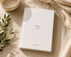 A white hardcover journal or book with the text 'Baptized 11/23' and 'THIO & CO.' printed on the cover, styled on a beige fabric surface with a ceramic bowl, green leaves, and a cream throw blanket.