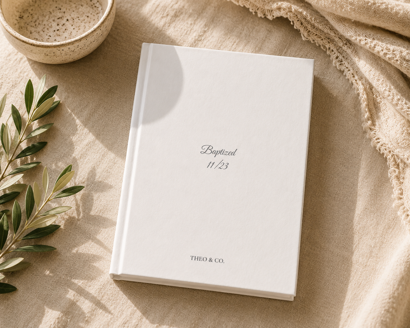 A white hardcover journal or book with the text 'Baptized 11/23' and 'THIO & CO.' printed on the cover, styled on a beige fabric surface with a ceramic bowl, green leaves, and a cream throw blanket.