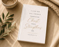 A white hardcover Bible or book with gold and black text reading 'I CAN DO ALL THINGS THROUGH Christ WHO Strengthens ME PHILIPPIANS 4:13', styled on a beige fabric surface with a small bowl, leafy branch, and knit textile nearby.