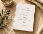 A white hardcover Bible or book with gold and black text reading 'I CAN DO ALL THINGS THROUGH Christ WHO Strengthens ME PHILIPPIANS 4:13', styled on a beige fabric surface with a small bowl, leafy branch, and knit textile nearby.
