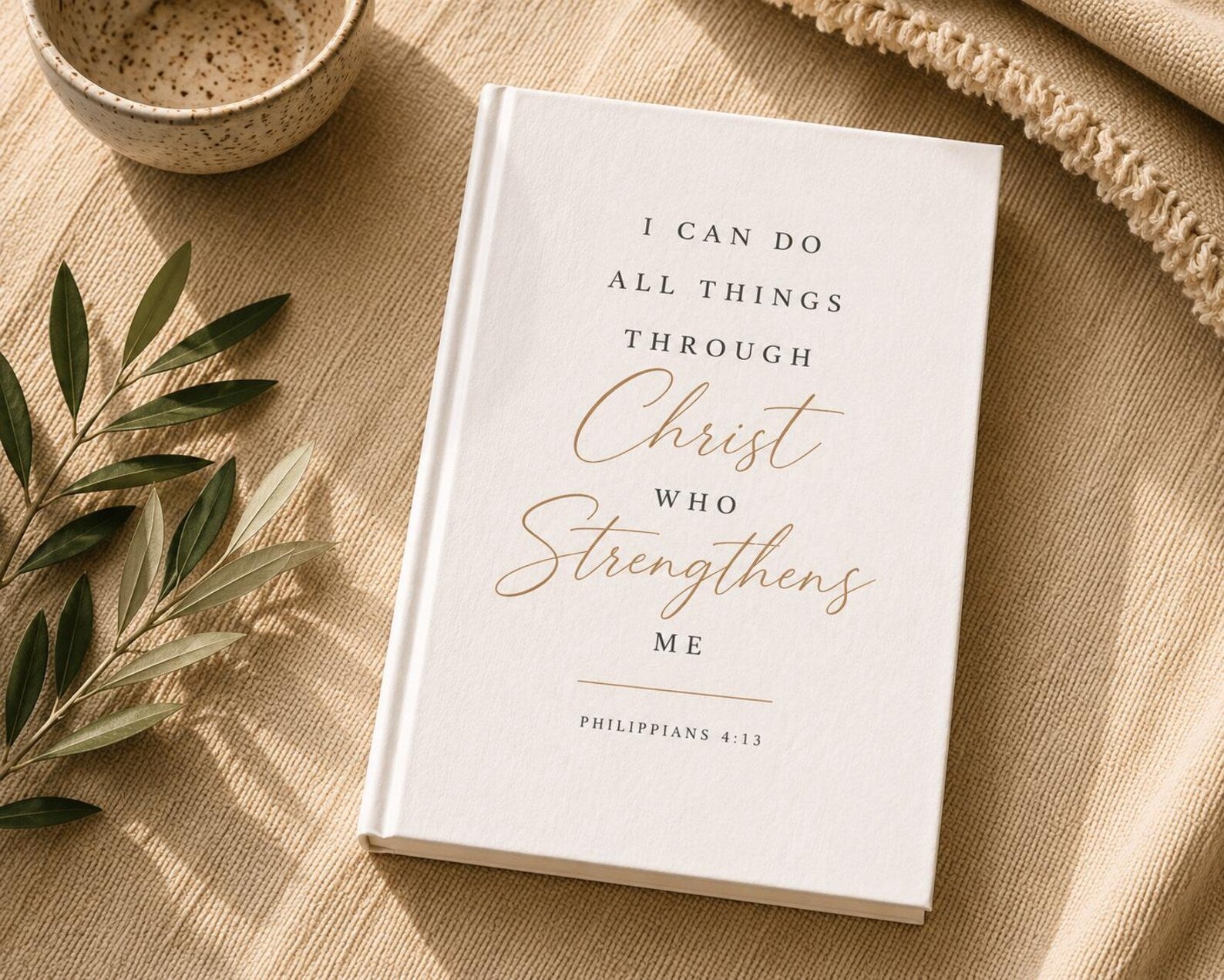 A white hardcover Bible or book with gold and black text reading 'I CAN DO ALL THINGS THROUGH Christ WHO Strengthens ME PHILIPPIANS 4:13', styled on a beige fabric surface with a small bowl, leafy branch, and knit textile nearby.