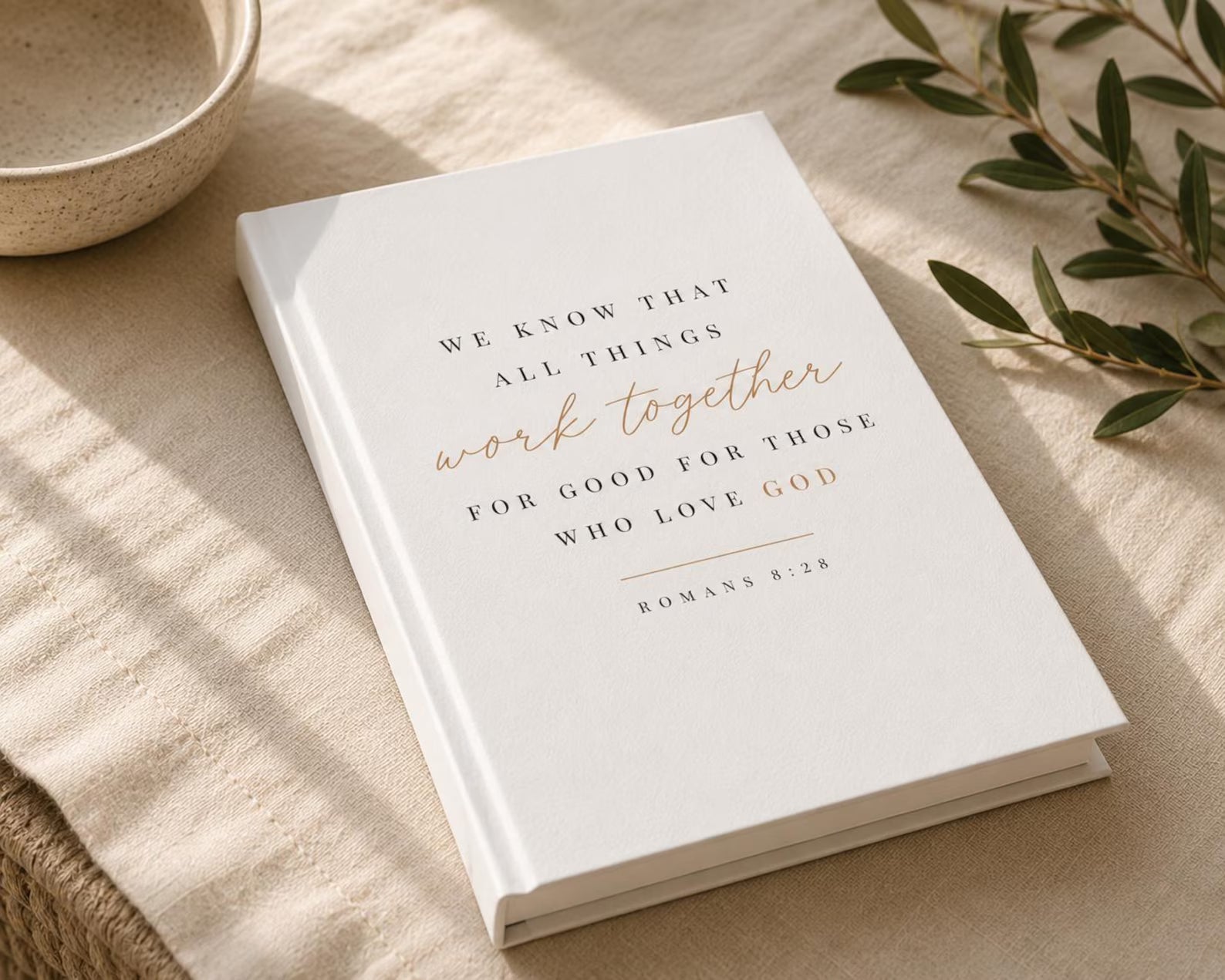 A white hardcover book with elegant gold and black typography reading 'WE KNOW THAT ALL THINGS work together FOR GOOD FOR THOSE WHO LOVE GOD ROMANS 8:28', styled on a beige linen surface with a ceramic bowl and green leaves.