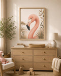 A framed art print of a pink flamingo hanging on a beige wall above a light wood dresser in a styled nursery or children's room. The room includes a matching pink plush toy, a white rocking chair, a lamp, and wooden toys.