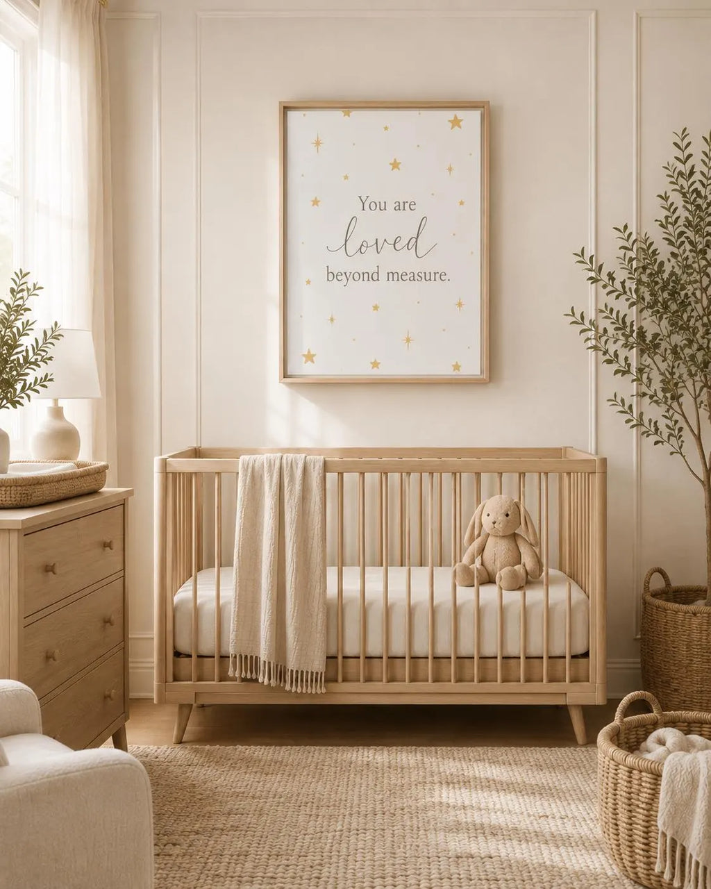 A styled nursery scene featuring a light wood crib with a cream blanket and stuffed bunny, beneath a framed wall print that reads 'You are loved beyond measure' with small gold stars. The room has warm neutral tones, a dresser, plants, and woven baskets.